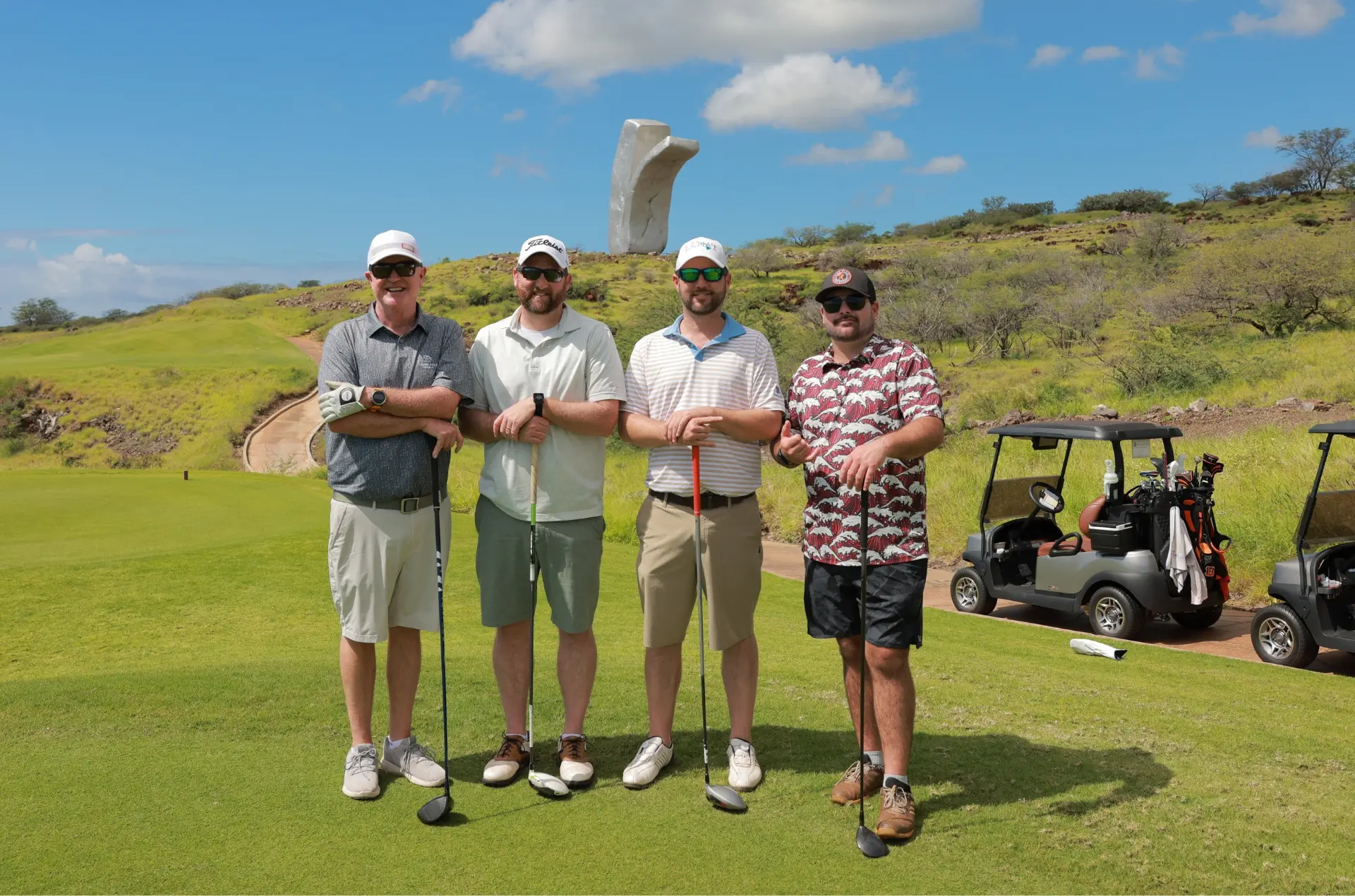 Photo of four men smiling and posing together on a golf course in Lanai. They are standing side by side, dressed in golf attire, with the lush landscape of Lanai in the background, capturing a joyful moment of friendship and leisure.