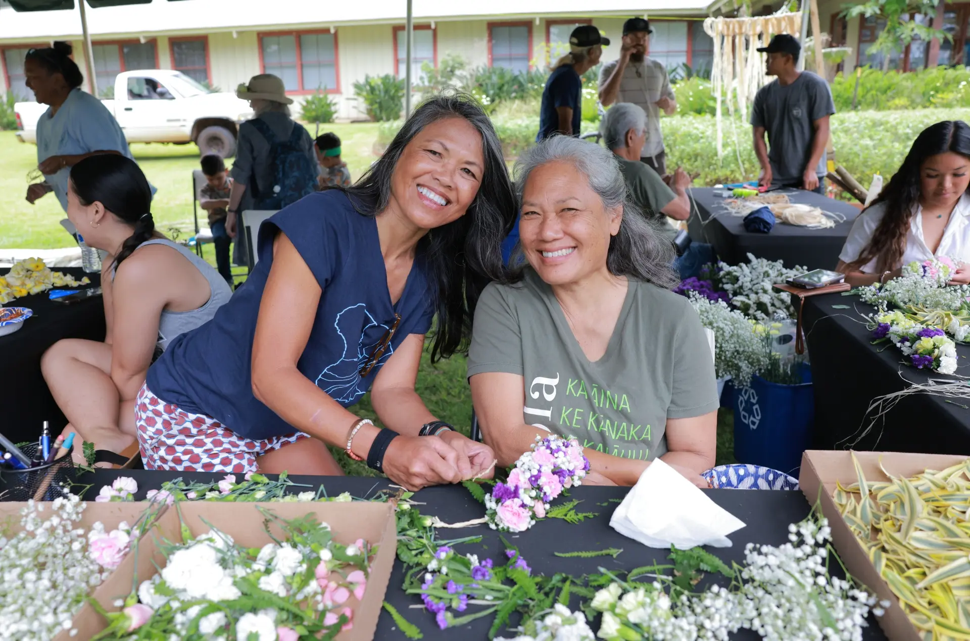 Photo of two women smiling at the camera while Kupulau is happening in the background.