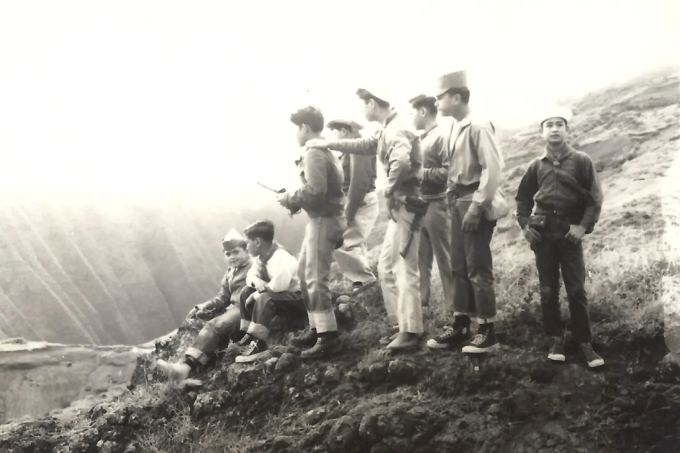 Black and white photo of a group of boys hiking through a natural landscape. The boys, dressed in outdoor gear, are walking along a trail, carrying backpacks and looking ahead, capturing a moment of adventure and camaraderie.