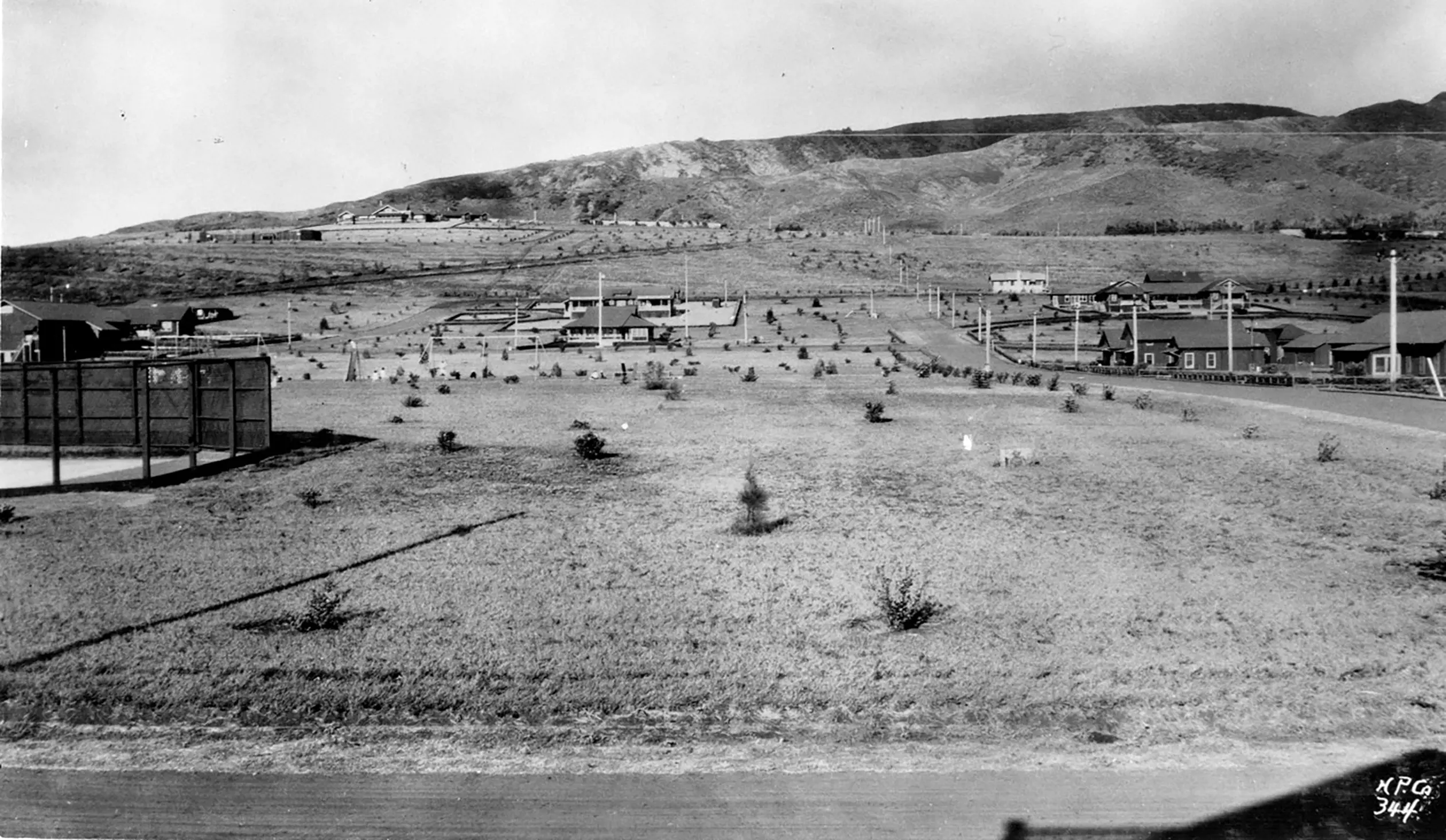 Black and white photo of Lanai City, showing an expansive view of land with scattered houses. The landscape is open and rural, with a few simple homes dotting the area, set against the backdrop of rolling hills and natural terrain, capturing the peaceful, undeveloped character of the region.