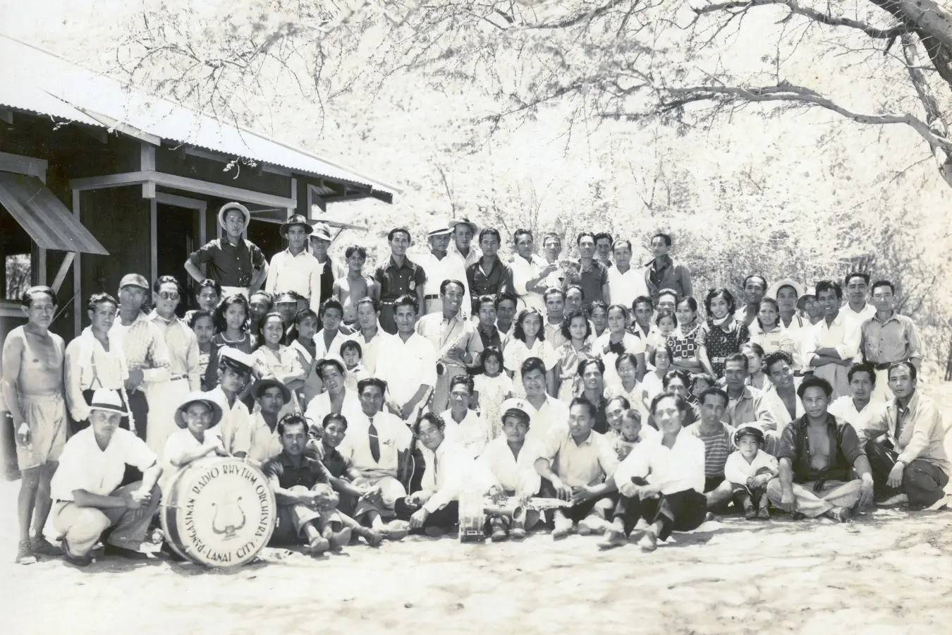 Black and white photo of the Lanai City Orchestra, with men and women gathered together for a group picture. The musicians are dressed in formal attire, standing side by side, showcasing a sense of unity and musical dedication.