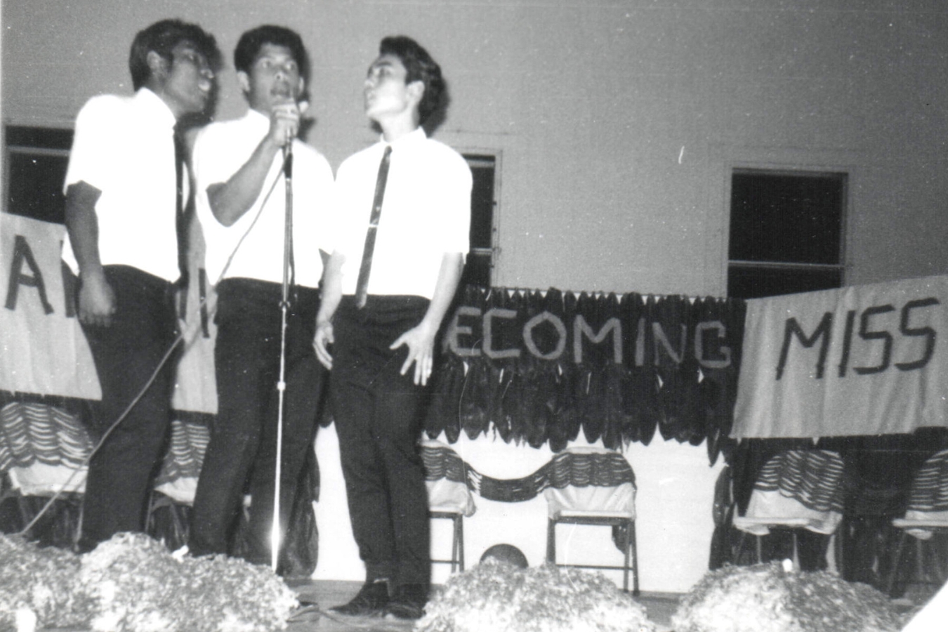 Three men wearing ties are positioned on stage, each with a microphone, likely addressing an audience.