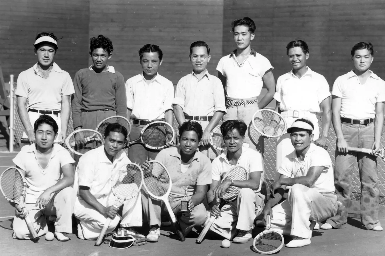 Black and white photo of a men's tennis team, all dressed in white uniforms, posing together for a team photo. The players stand side by side, holding their rackets with pride.