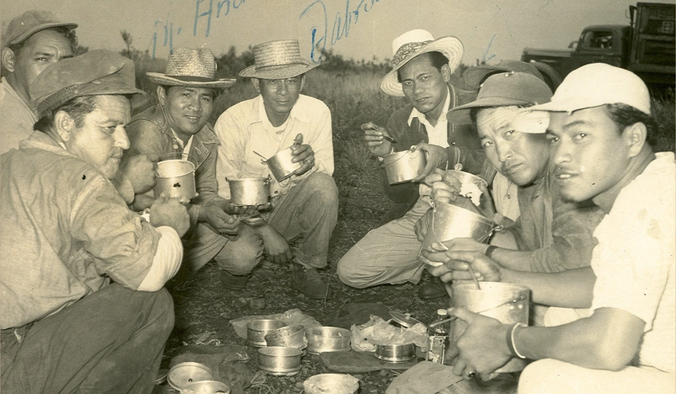 Historical image of ranching men sitting in a half circle eating out of metal canteen bowls.